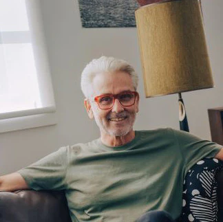An elderly man with white hair and glasses, smiling, sitting on a couch in a living room.