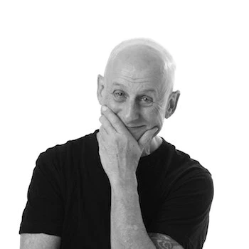 Black and white photo of a bald elderly man in a black shirt, smiling and resting his chin on his hand.